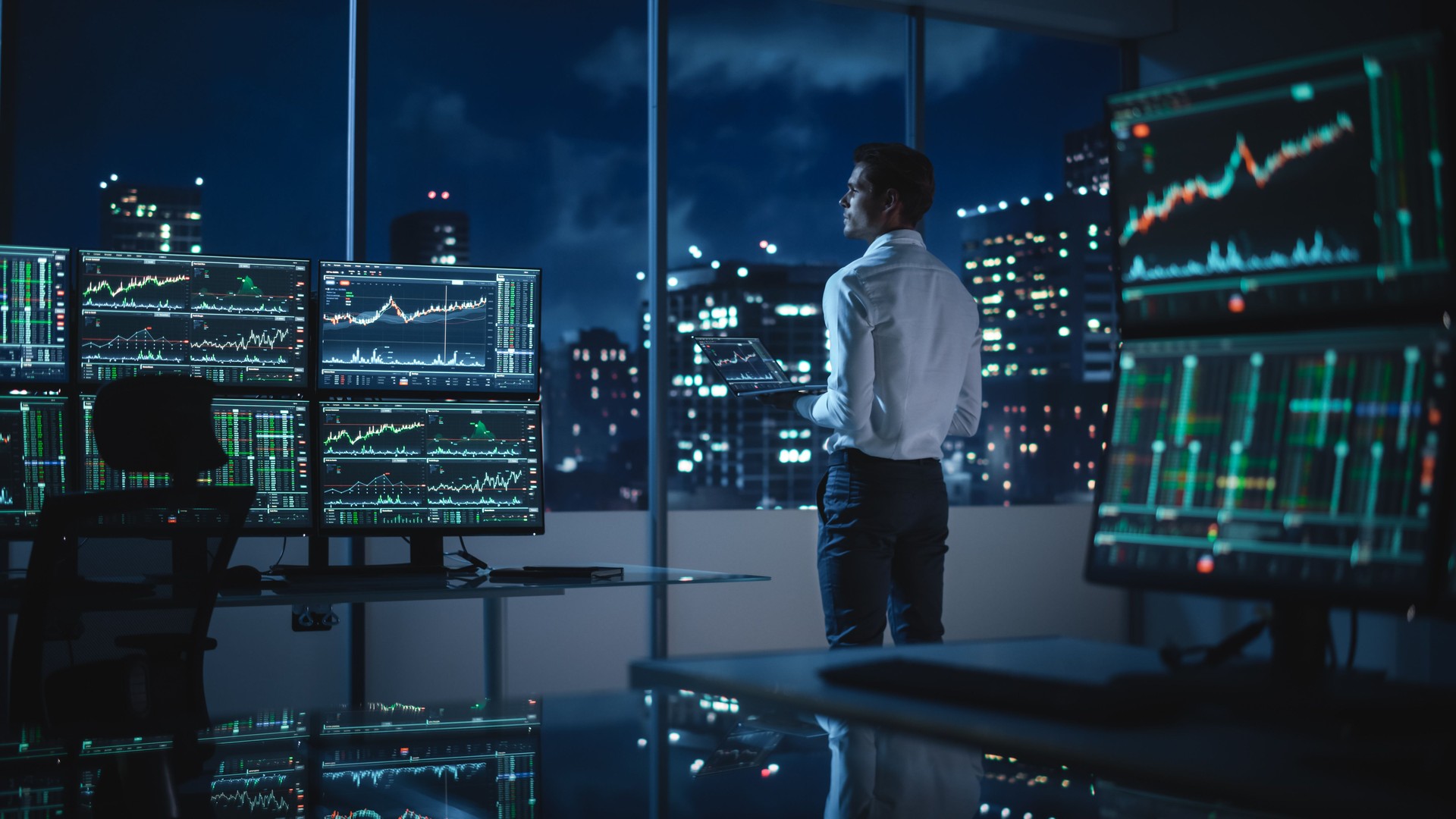 Financial Analyst Using Laptop Computer, Standing Next to Multi-Monitor Workstation with Real-Time Stocks, Commodities and Exchange Market Charts. Businessman Working in Broker Agency Office.